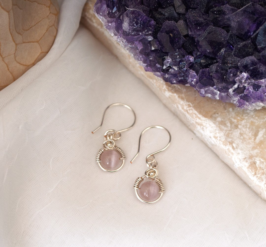 Handmade pink cat's eye earrings with 6mm light pink cat's eye bead in silver-plated wire halo with wire-wrapped rose detail, displayed on white linen surface beside raw amethyst crystal cluster