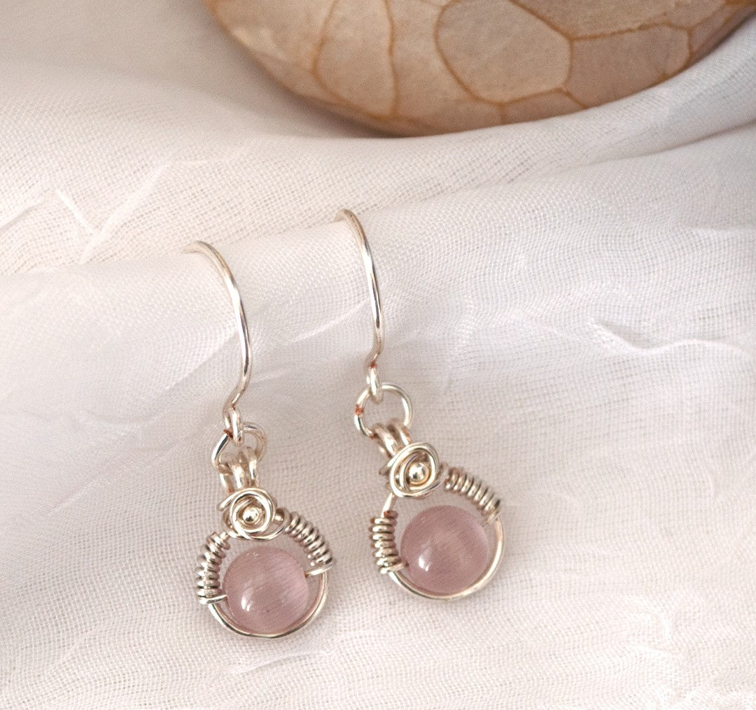 Close-up of handmade pink cat's eye earrings with silver-plated wire halo and wire-wrapped rose with silver bead center, displayed on white linen with petrified wood