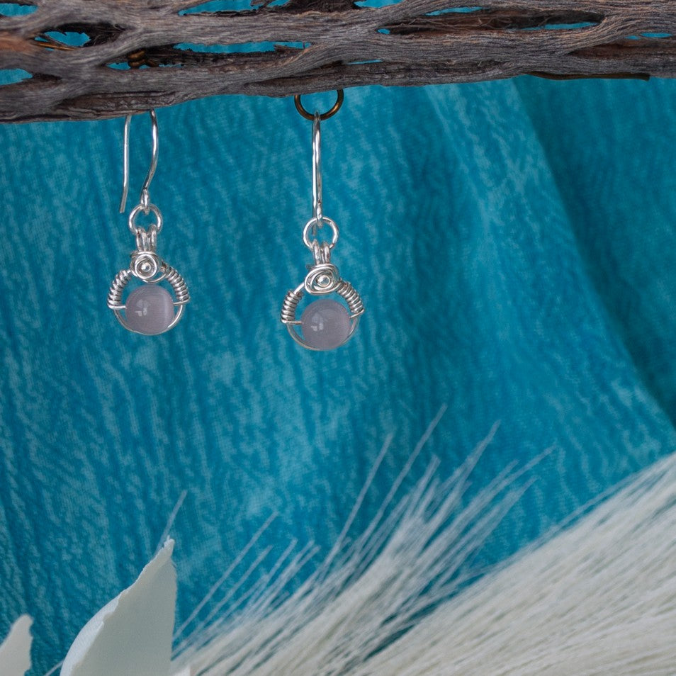 Pink cat's eye earrings with handcrafted silver wire halo and wire-wrapped rose detail hanging from driftwood with teal water background and pampas grass