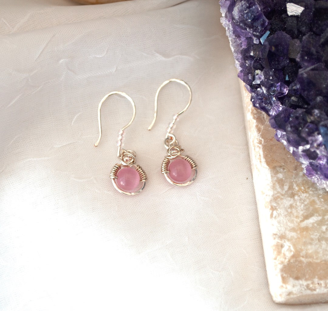Pink cat's eye earrings with silver-plated wire halo and iridescent seed bead accents on hammered ear wires, displayed on white linen surface beside raw amethyst crystal cluster