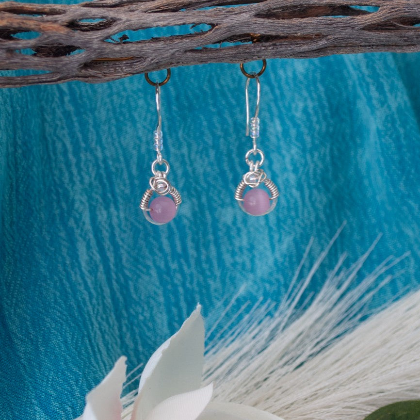 Handmade pink cat's eye earrings with silver wire halo and Miyuki seed bead accents hanging from driftwood with teal water background and white magnolia flower