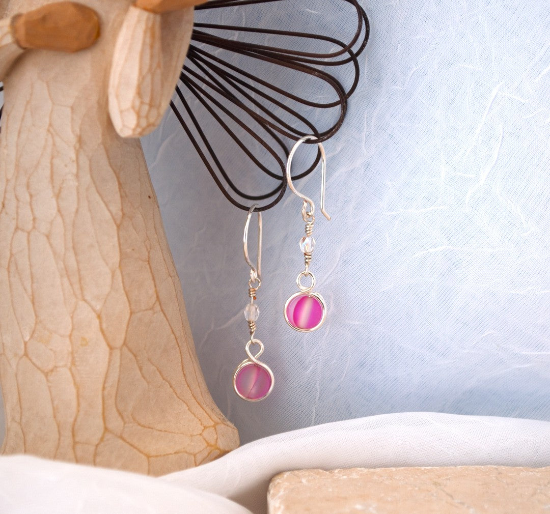 Handmade pink mermaid glass earrings with silver-plated double wire wrap and fire polished Czech bead accent displayed on Willow Tree angel figurine against blue-gray background with travertine