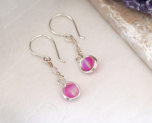 Handmade pink mermaid glass earrings with 6mm double wire-wrapped pink mermaid glass bead and 3mm fire polished Czech bead accent on silver-plated ear wires, displayed on white linen surface beside raw amethyst crystal cluster