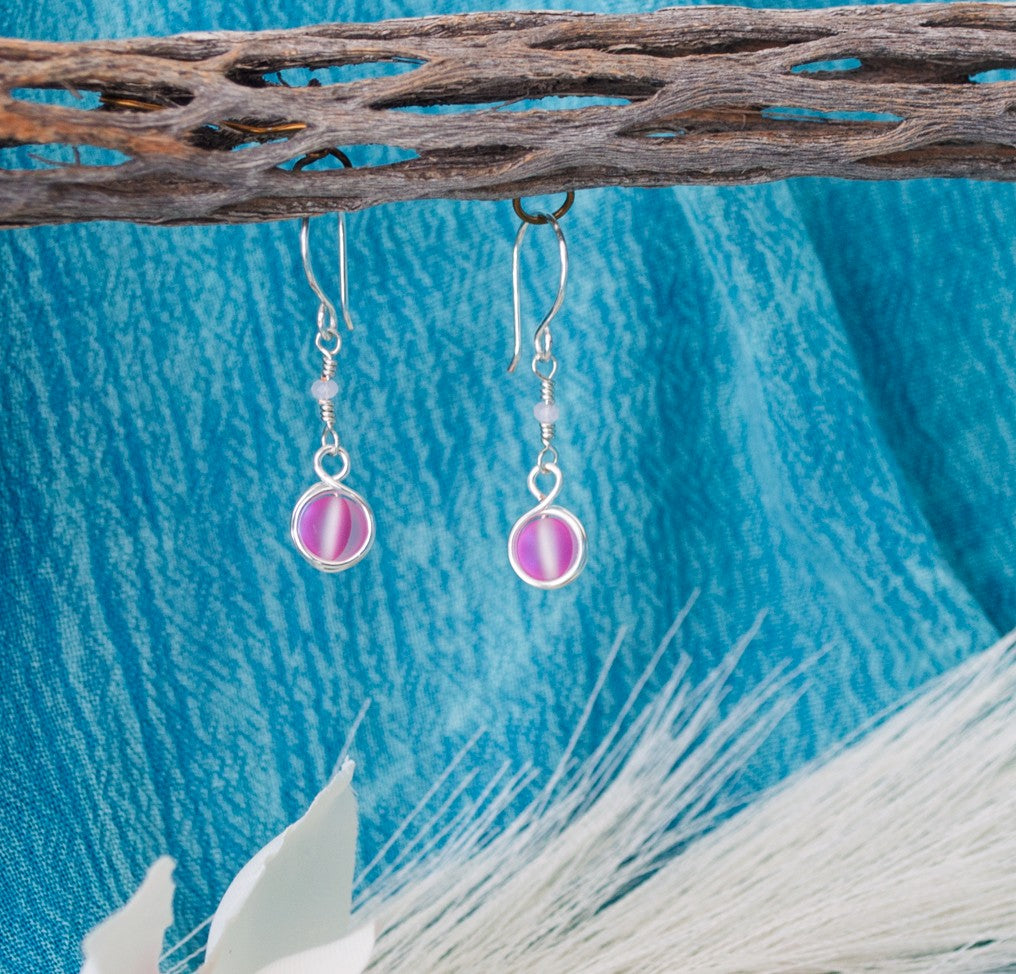 Pink mermaid glass earrings with double wire-wrapped bead and faceted crystal rondelle accent hanging from driftwood with teal water background and white magnolia flower