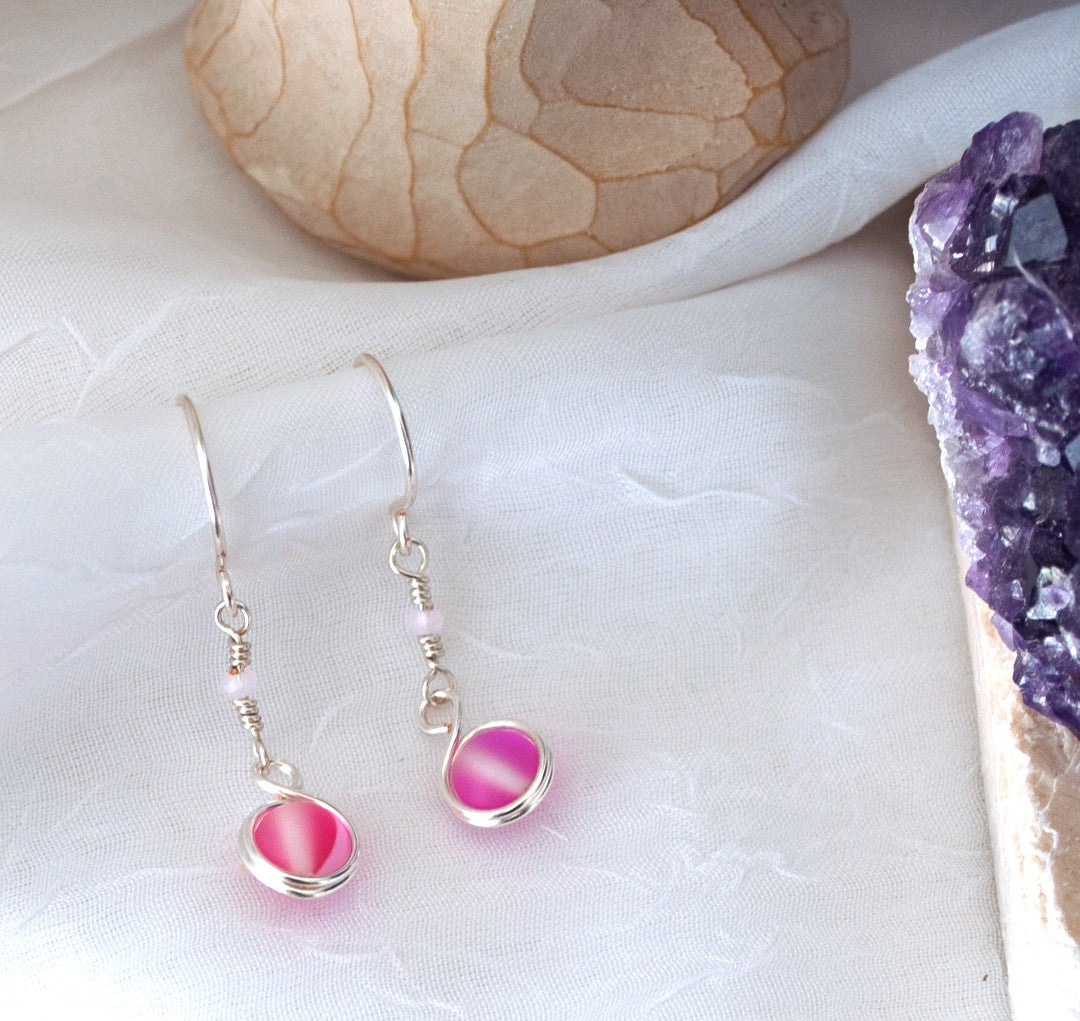 Handmade pink mermaid glass earrings with silver-plated double wire wrap and light pink crystal rondelle accent displayed on white linen drape beside raw amethyst crystal cluster