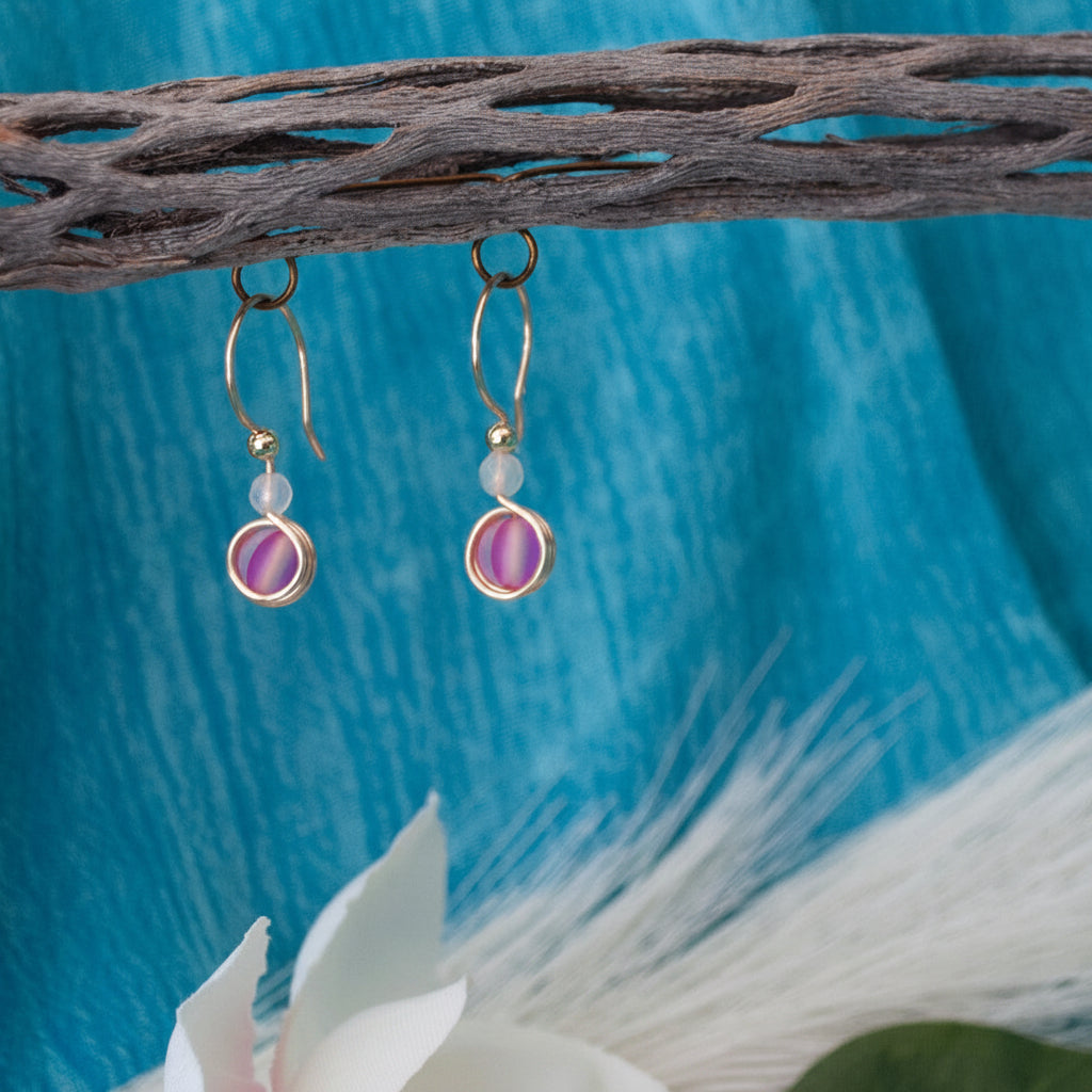 A pair of brass French hook dangle earrings with white faceted agate beads and round vivid fuchsia beads, hanging from a rustic branch against a bright blue textured background with white decorative elements.