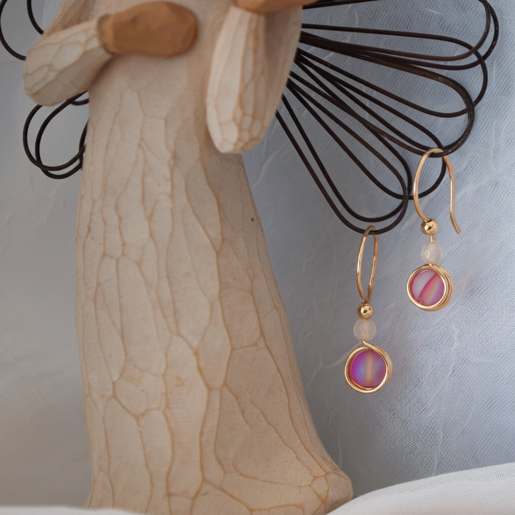 Brass drop earrings with vivid fuchsia round beads and small white agate beads, displayed on a decorative stand against a pale blue fabric background.
