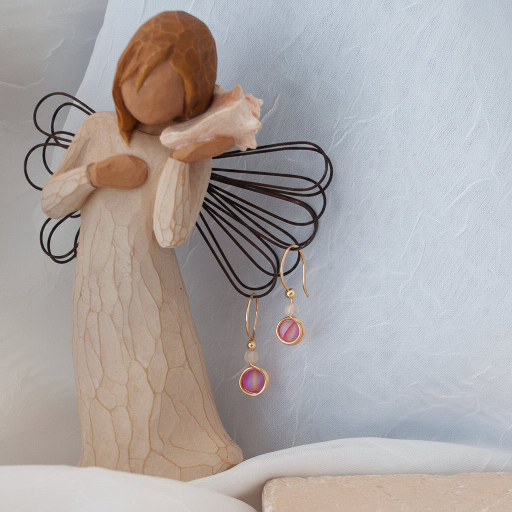 Brass dangle earrings with round vivid fuchsia glass beads and white agate beads, shown hanging from a decorative angel figurine against a soft light blue textured backdrop.