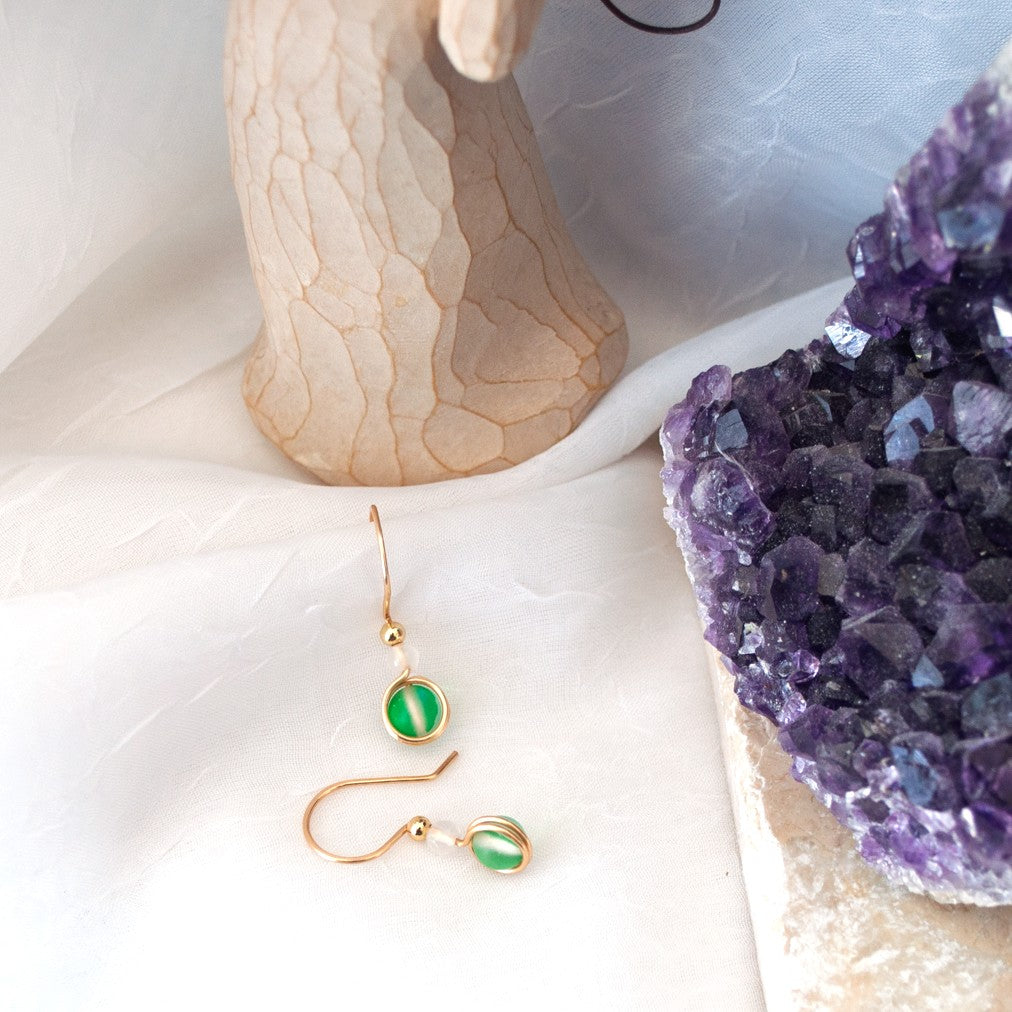 Green mermaid glass drop earrings with white faceted agate beads and gold-tone brass wire arranged on a white surface beside a carved angel and purple amethyst.