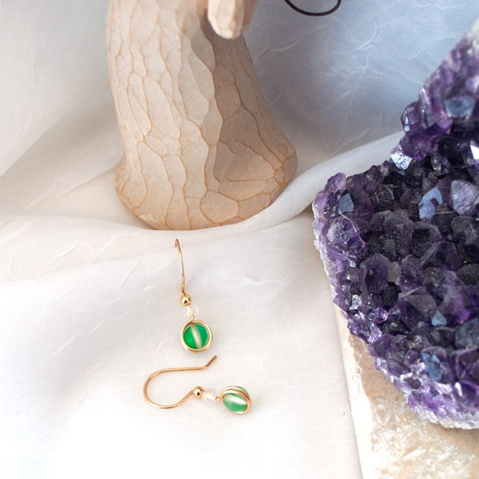Green mermaid glass drop earrings with white faceted agate beads and gold-tone brass wire arranged on a white surface beside a carved angel and purple amethyst.
