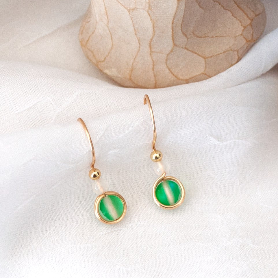 Pair of green mermaid glass drop earrings with white faceted agate beads and gold-tone brass wire displayed on a white surface beside a carved angel and purple amethyst