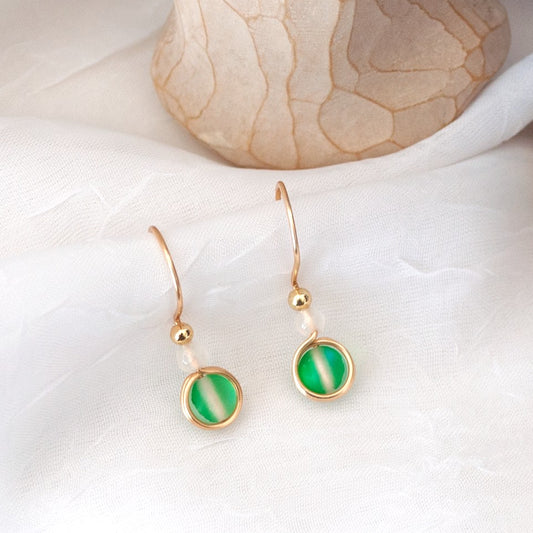 Pair of green mermaid glass drop earrings with white faceted agate beads and gold-tone brass wire displayed on a white surface beside a carved angel and purple amethyst