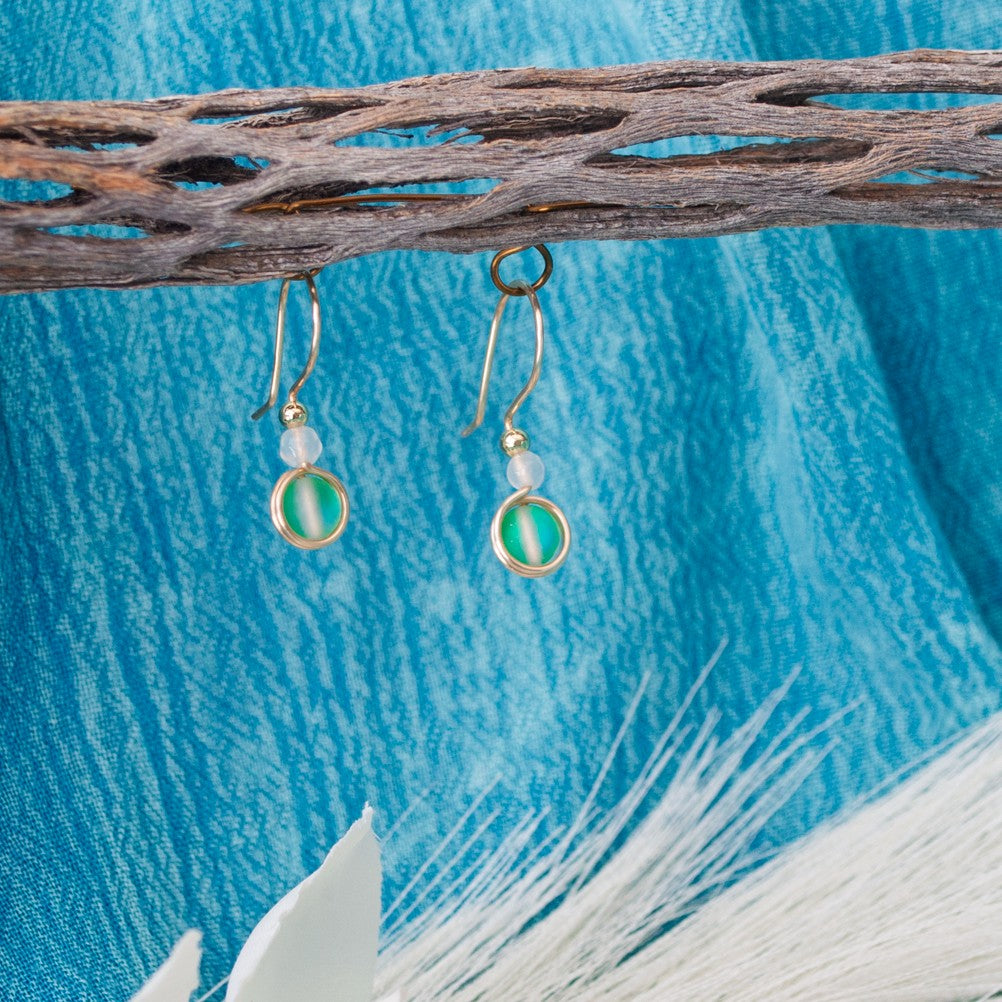 Pair of green mermaid glass drop earrings with white faceted agate beads hanging from a rustic branch against teal textured fabric