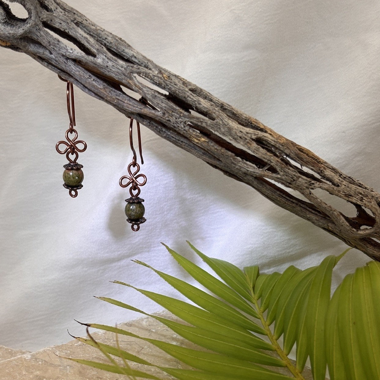 Handmade unakite gemstone drop earrings with antique copper clover charm and handcrafted copper ear wires, hanging from driftwood with tropical palm leaves.
