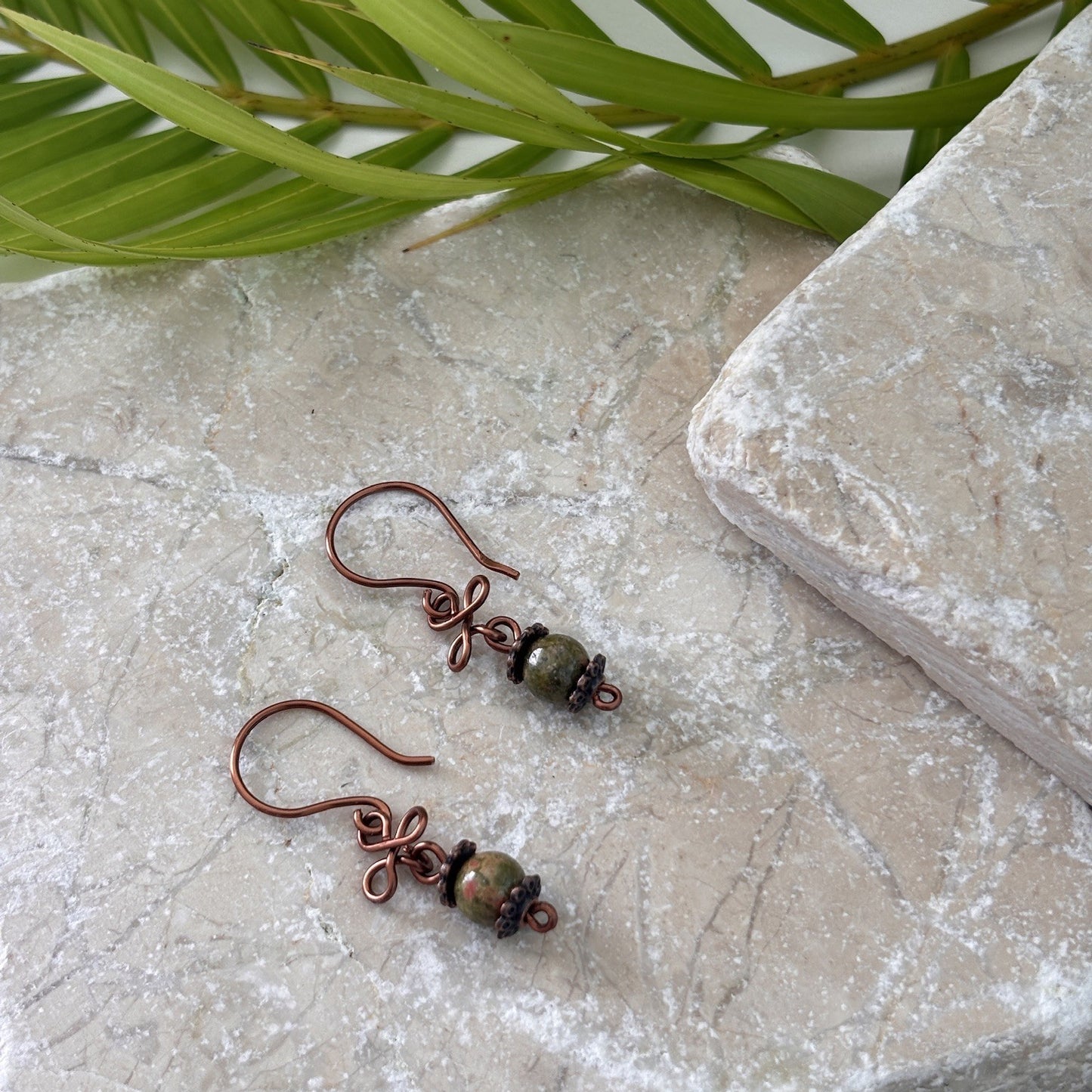 Pair of handmade unakite gemstone drop earrings with antique copper clover charm and handcrafted copper ear wires, displayed on travertine stone with tropical palm leaves.