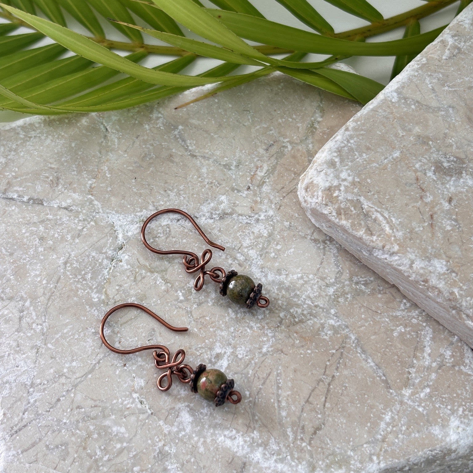 Pair of handmade unakite gemstone drop earrings with antique copper clover charm and handcrafted copper ear wires, displayed on travertine stone with tropical palm leaves.