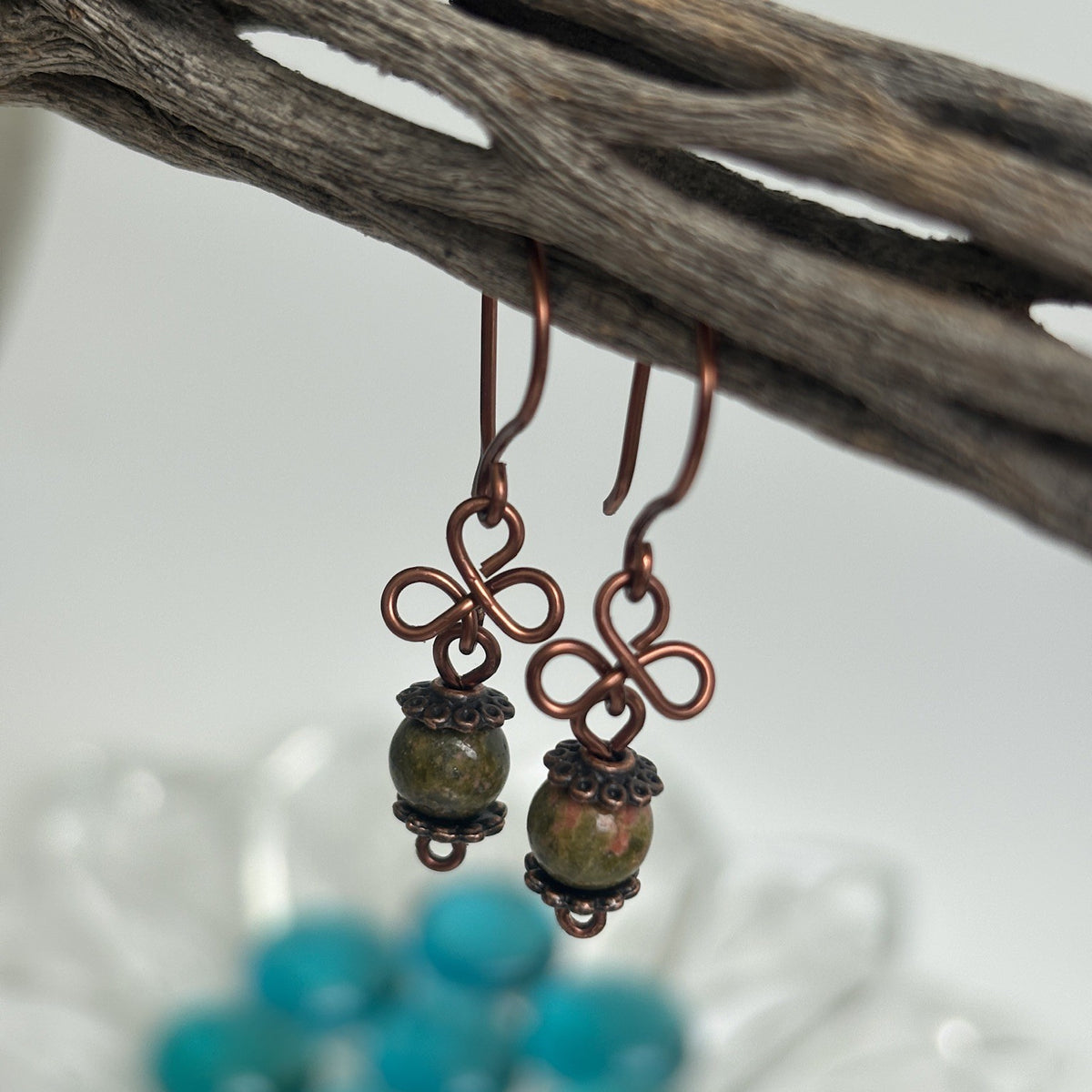 Handmade unakite gemstone drop earrings with antique copper clover charm and copper bead caps, hanging from driftwood against a soft neutral background