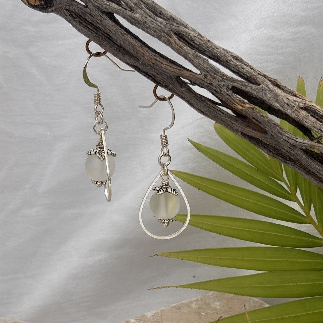 Handmade frosted mermaid glass drop earrings in silver-tone teardrop frame with leaf bead cap and daisy spacer, hanging from driftwood with tropical palm leaves.