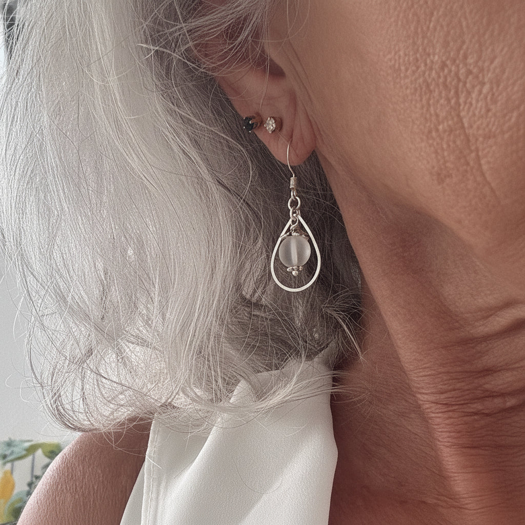 Handmade frosted mermaid glass drop earring in silver-tone teardrop frame shown worn on model with silver hair.

