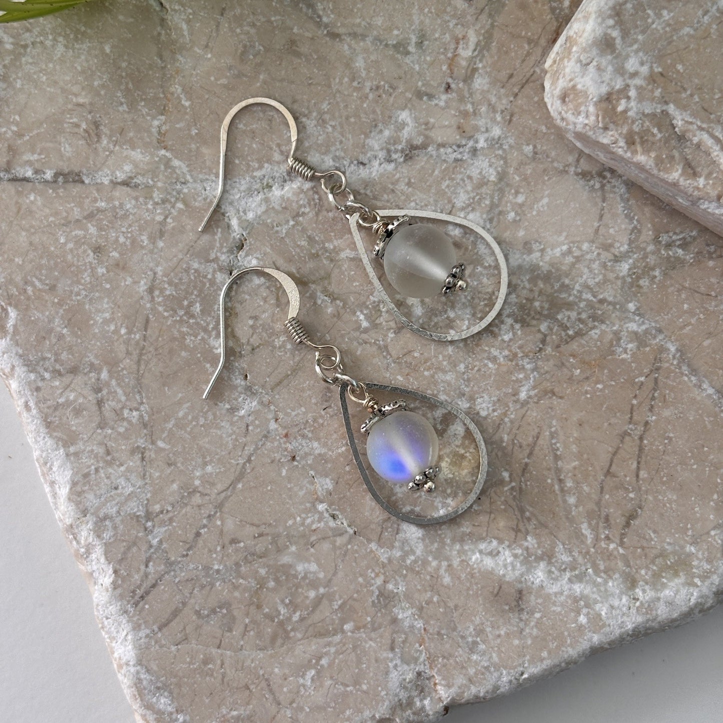Pair of handmade frosted mermaid glass drop earrings in silver-tone teardrop frame displayed on travertine stone, showing blue iridescent flash in the mermaid glass bead.