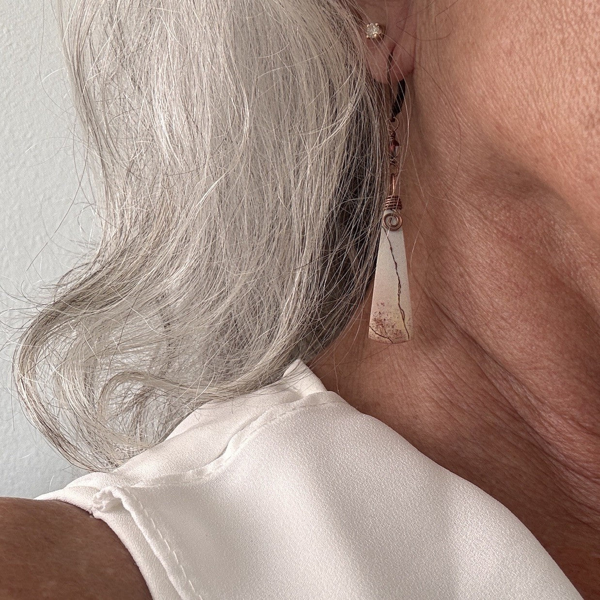 Handmade porcelain jasper drop earring with antique copper wire wrap shown worn on model with silver hair and white satin blouse.

