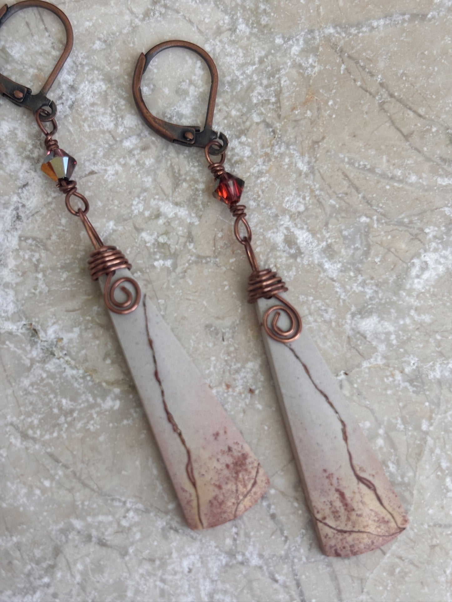 Pair of handmade porcelain jasper drop earrings with antique copper wire wrap and Swarovski crystal charms displayed on travertine stone.