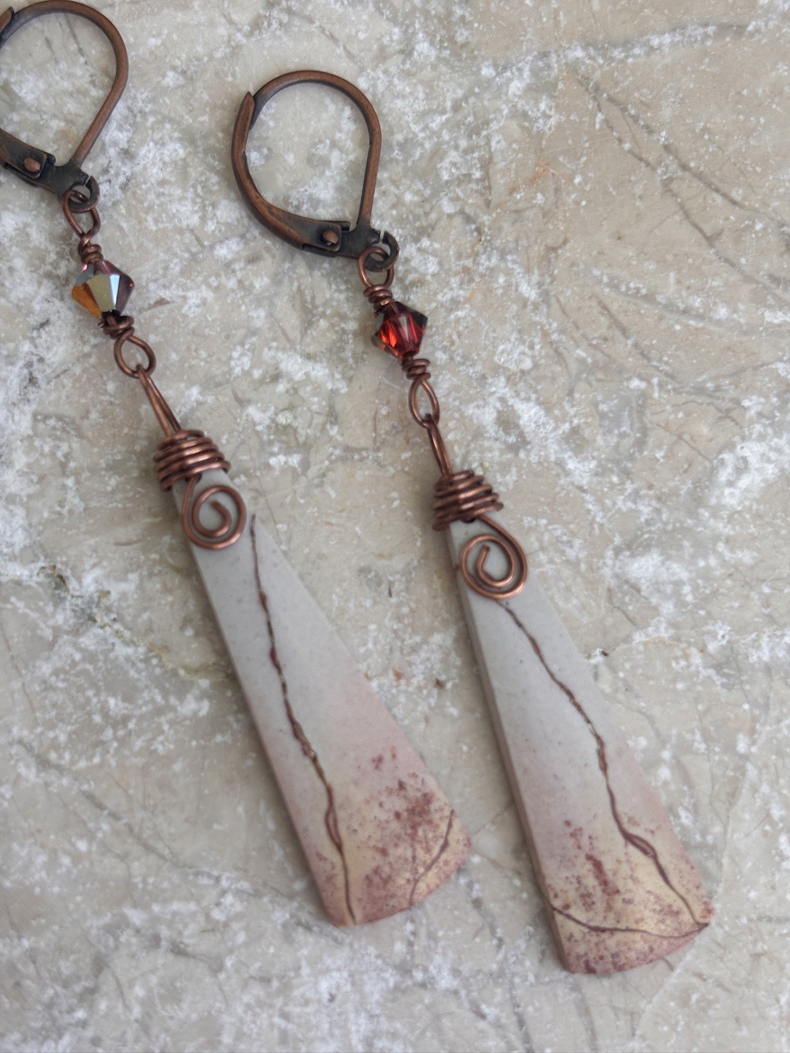 Pair of handmade porcelain jasper drop earrings with antique copper wire wrap and Swarovski crystal charms displayed on travertine stone.