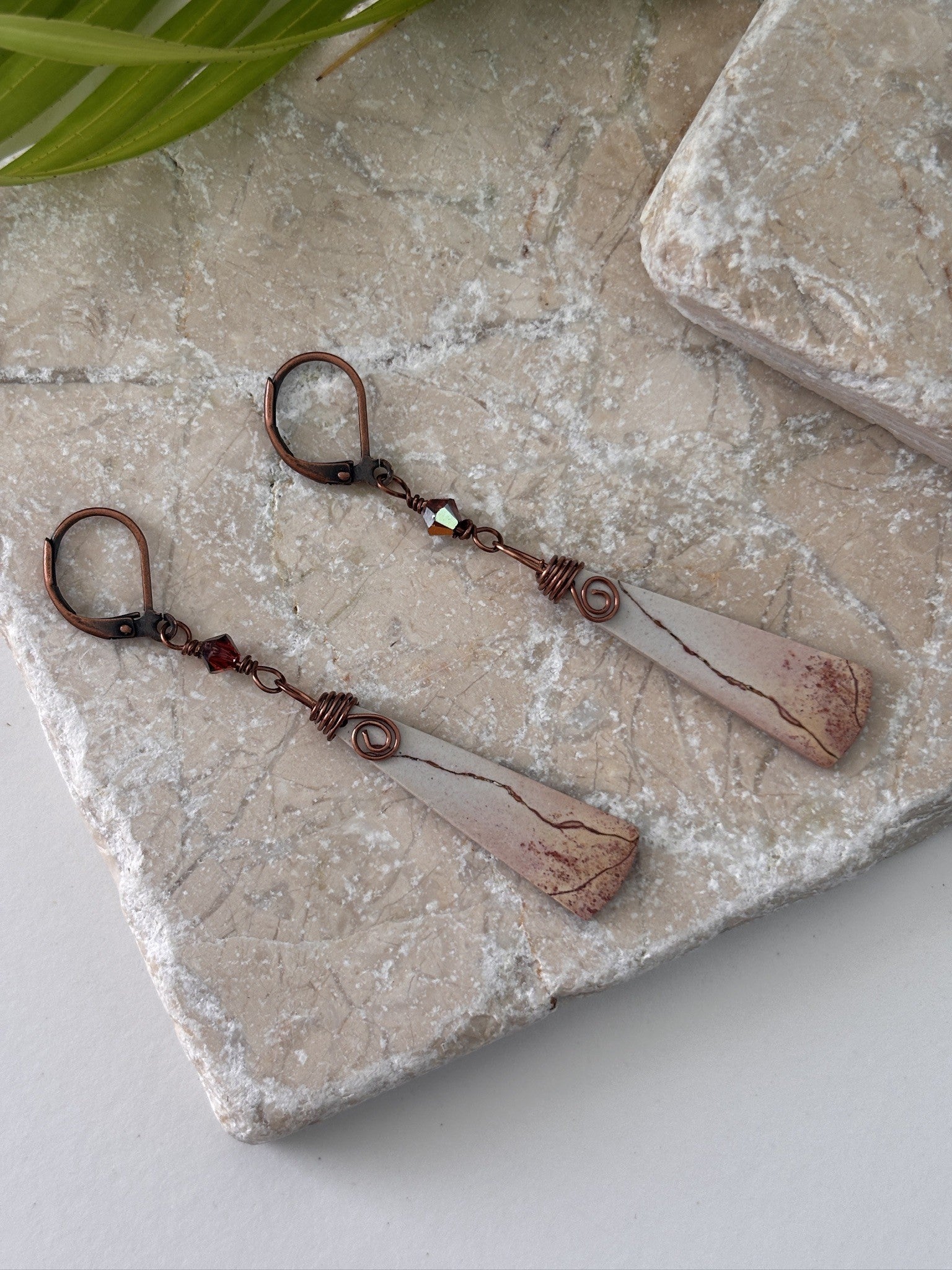 Handmade porcelain jasper drop earrings with antique copper wire wrap displayed on travertine stone tiles with tropical palm leaf accent.