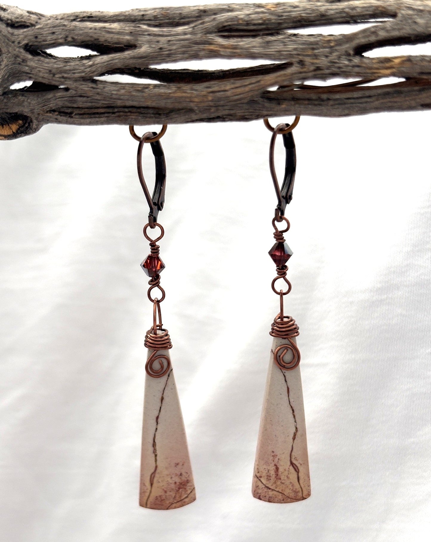 Pair of handmade porcelain jasper drop earrings with antique copper leverback ear wires, garnet Swarovski bicone crystal charms, and spiral wire wrap, hanging from driftwood.