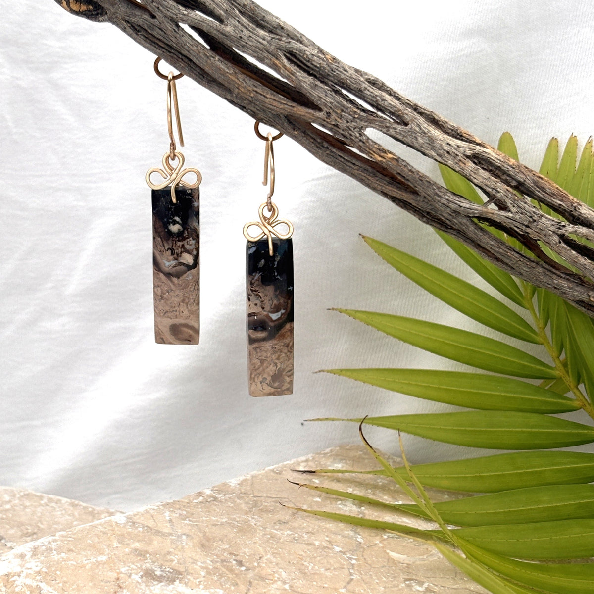 Petrified palm earrings with brass clover bail hanging from driftwood with palm leaf and white linen background