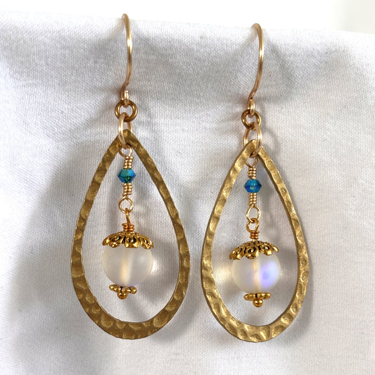 Handmade mermaid glass earrings with 20x35mm hammered brass teardrop frame, wire-wrapped drop with 8mm matte clear iridescent mermaid glass bead, gold-tone bead caps, and 3mm blue-green Swarovski bicone, displayed on white linen background