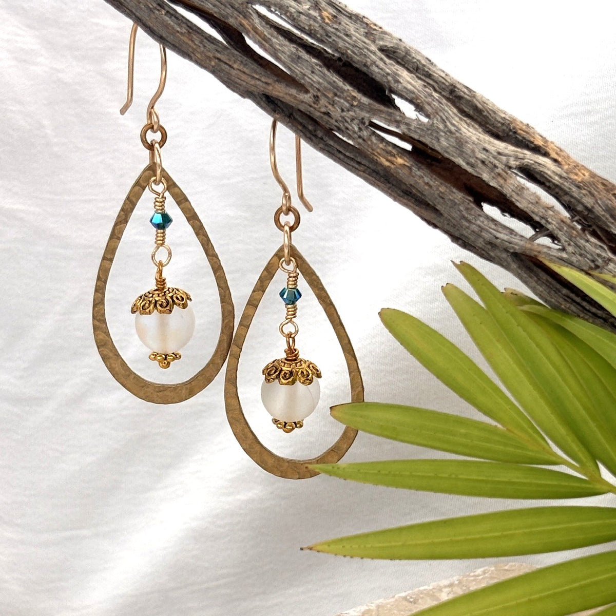 Mermaid glass earrings with hammered brass teardrop frame and blue-green Swarovski bicone hanging from driftwood with palm leaf and white linen background