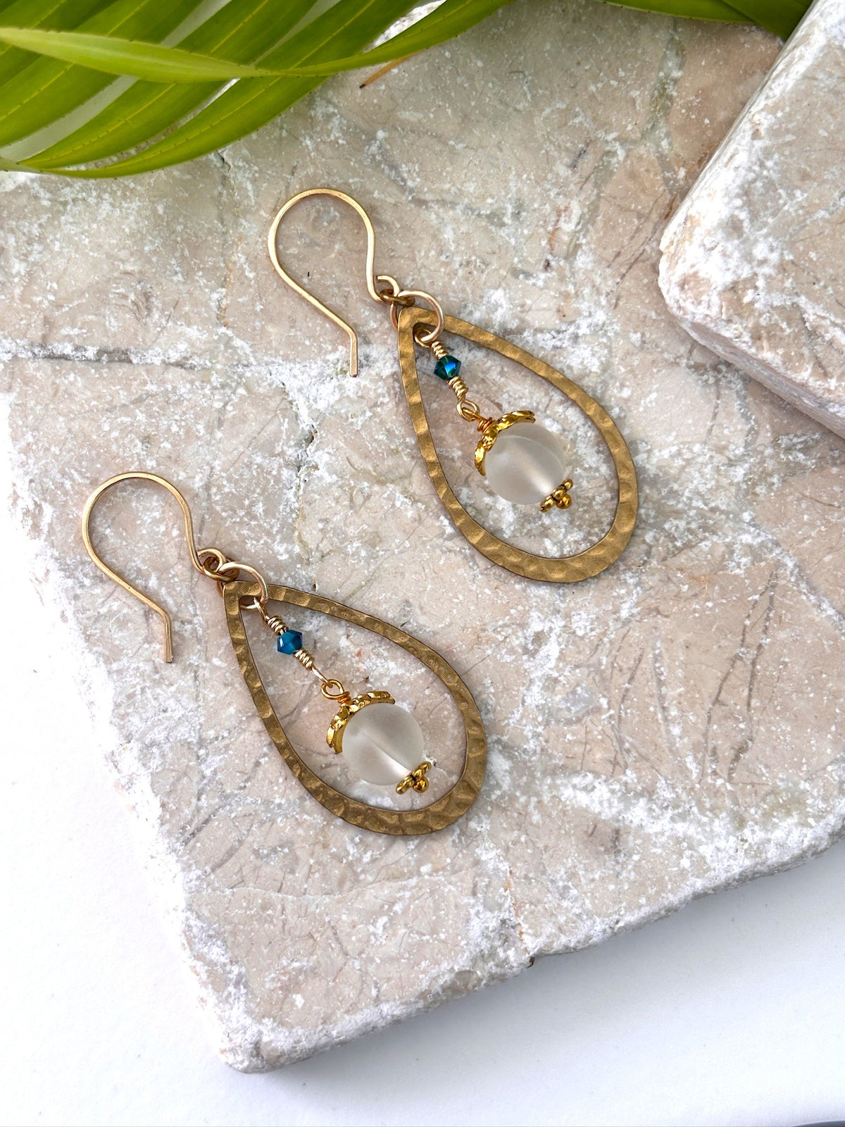 Pair of mermaid glass earrings with hammered brass teardrop frame and wire-wrapped mermaid glass drop displayed on travertine marble surface with palm leaf
