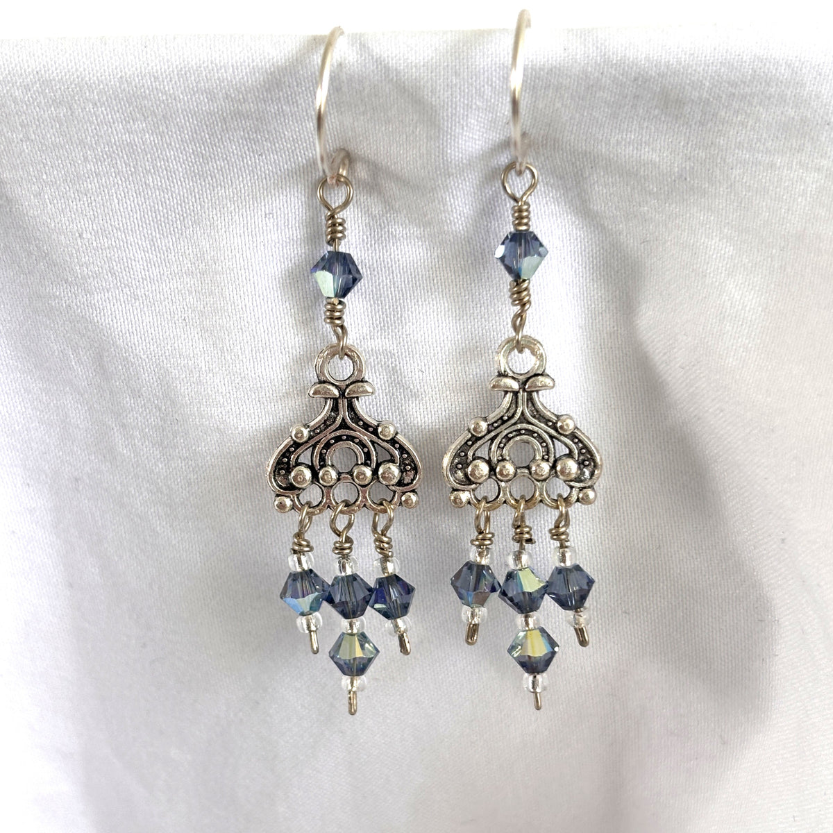 Handmade blue iridescent crystal chandelier earrings with ornate silver-tone chandelier component, three Swarovski bicone dangles with silver-lined seed bead accents, and wire-wrapped Swarovski bicone on silver-plated ear wires, displayed on white linen background