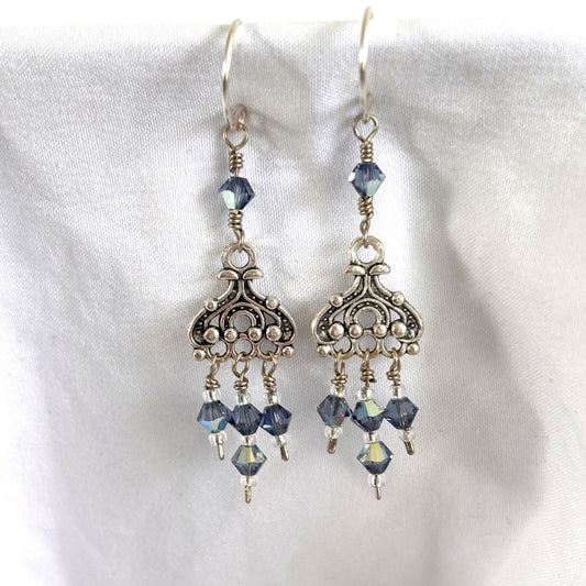 Handmade blue iridescent crystal chandelier earrings with ornate silver-tone chandelier component, three Swarovski bicone dangles with silver-lined seed bead accents, and wire-wrapped Swarovski bicone on silver-plated ear wires, displayed on white linen background