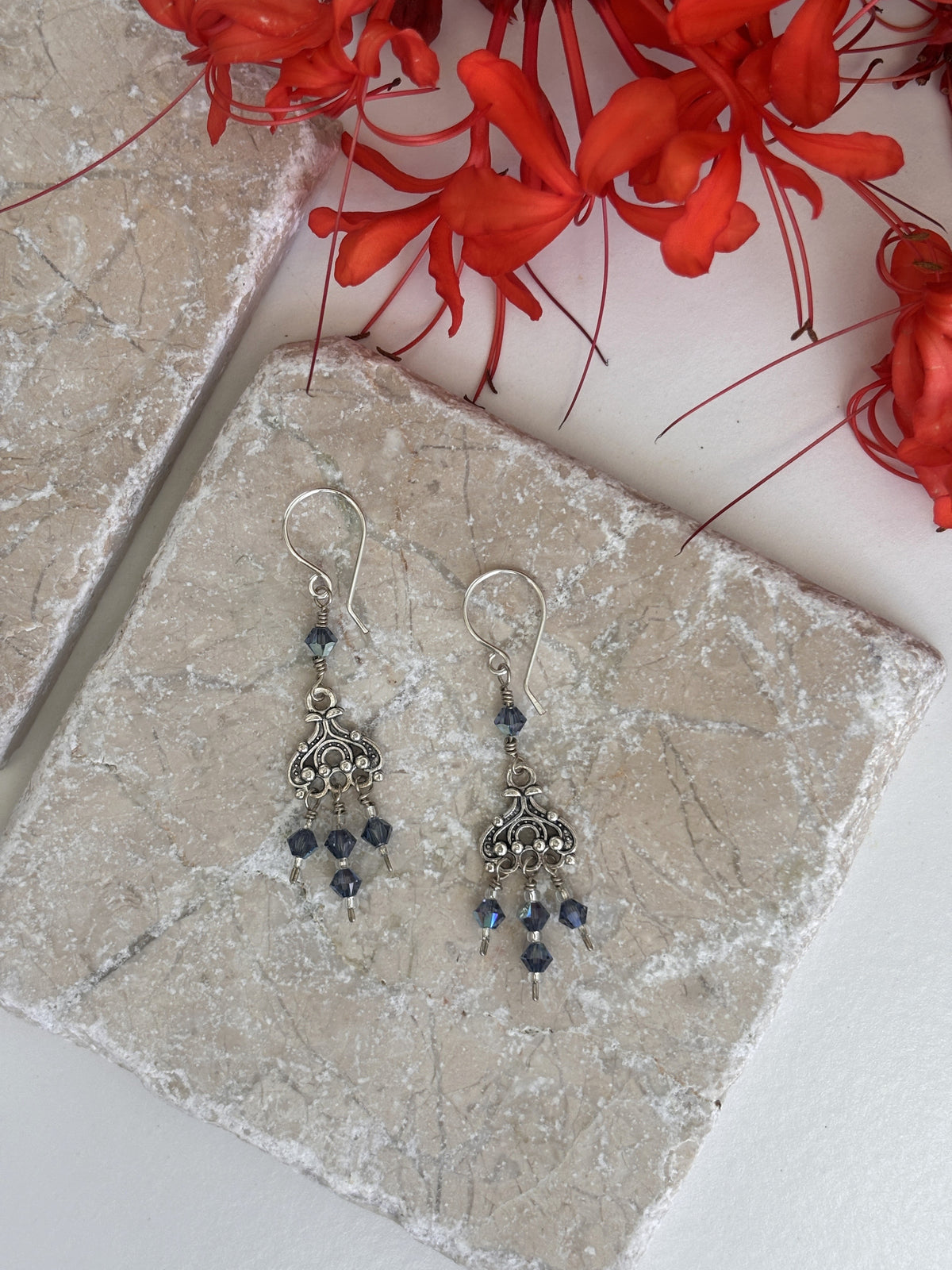 Handmade blue iridescent crystal chandelier earrings with ornate silver chandelier component and Swarovski bicone dangles displayed on travertine surface with red spider lily flowers