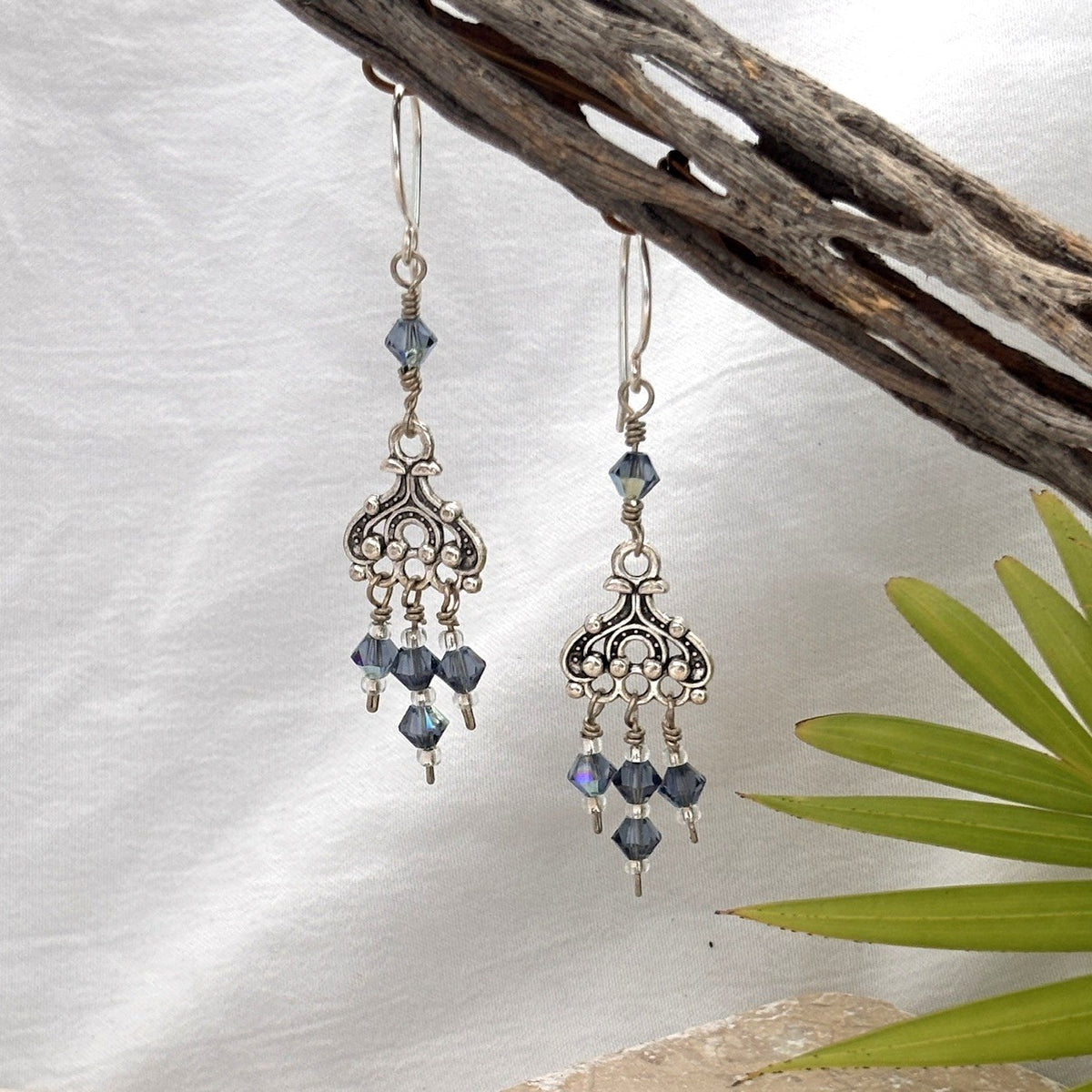 	Blue iridescent Swarovski crystal chandelier earrings with ornate silver-tone chandelier component and three crystal dangles hanging from driftwood with white linen background and palm frond