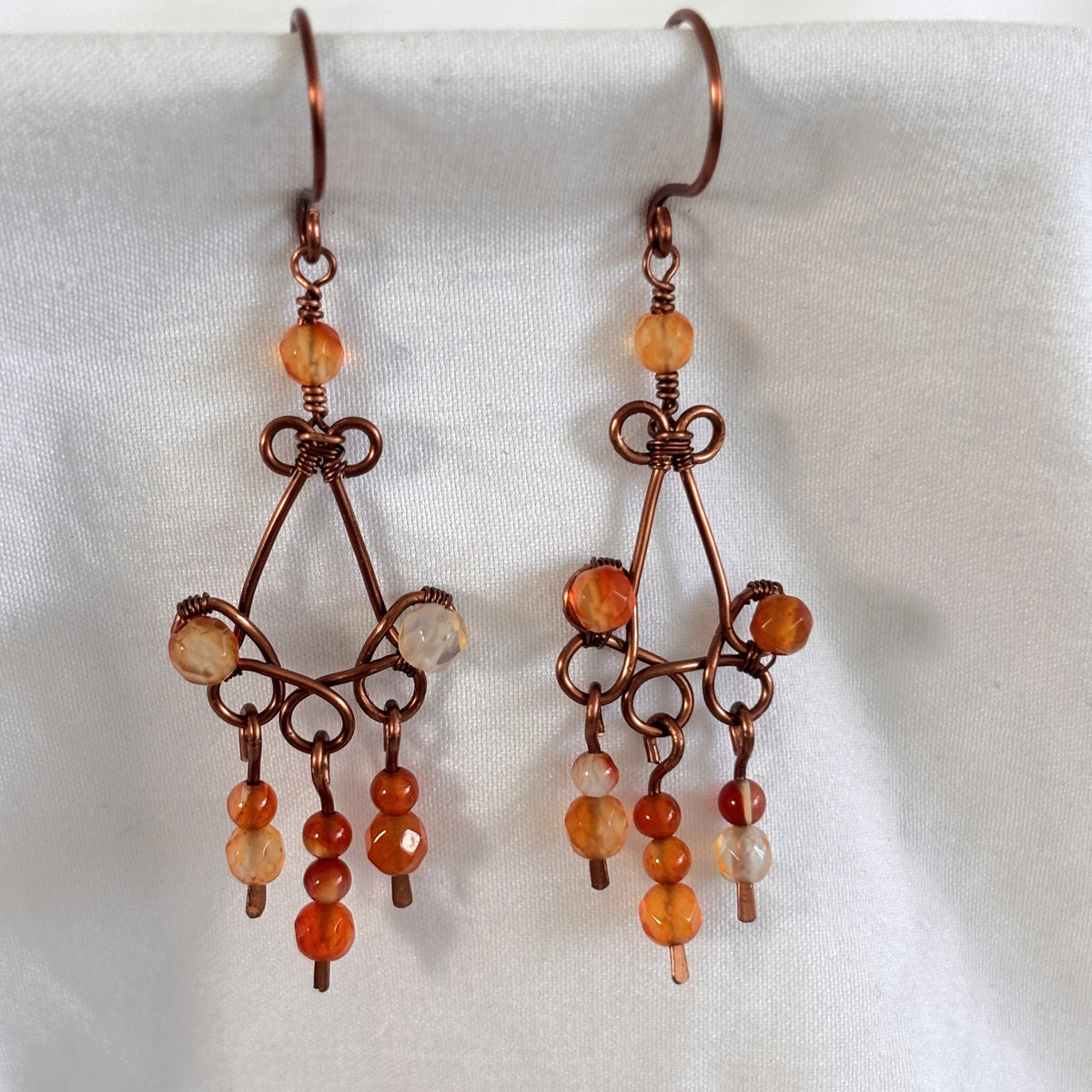 Handmade carnelian earrings with antique copper chandelier frame and three beaded paddles holding faceted and smooth carnelian beads in peach to amber-orange, displayed against white linen background