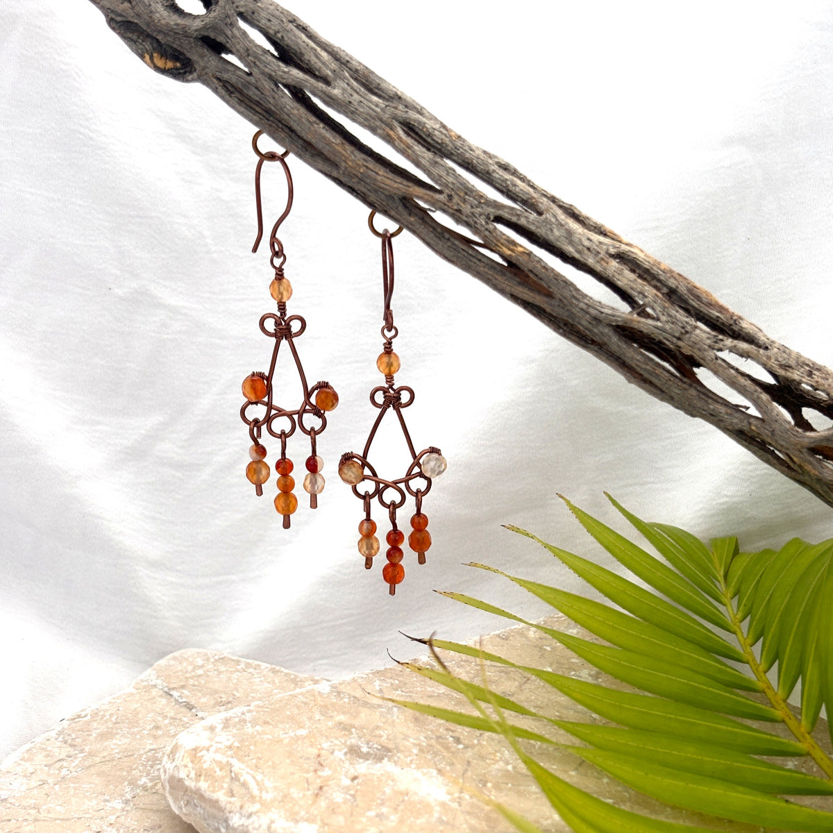 Carnelian earrings with antique copper chandelier frame and faceted carnelian bead paddles hanging from driftwood against white linen background with palm frond and travertine