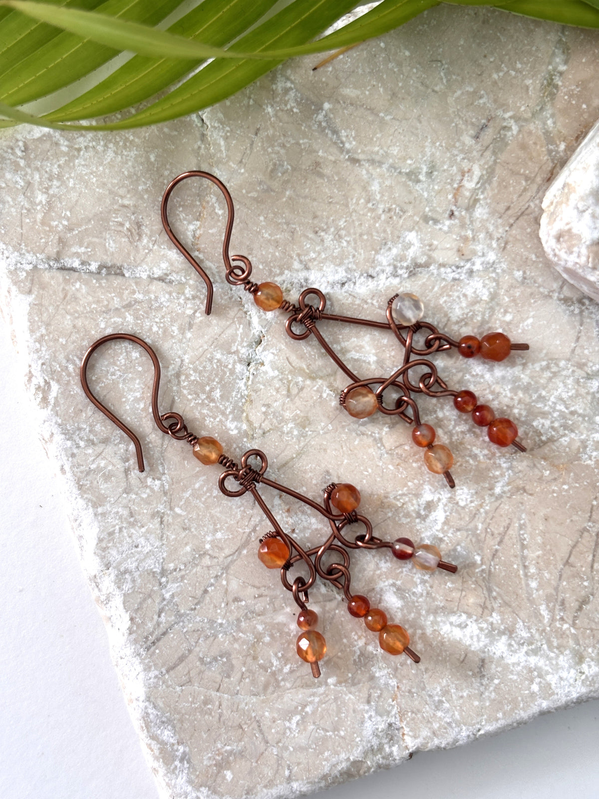Handmade antique copper carnelian chandelier earrings with faceted 4mm and smooth 3mm carnelian bead paddles displayed on travertine surface with palm frond and quartz crystal