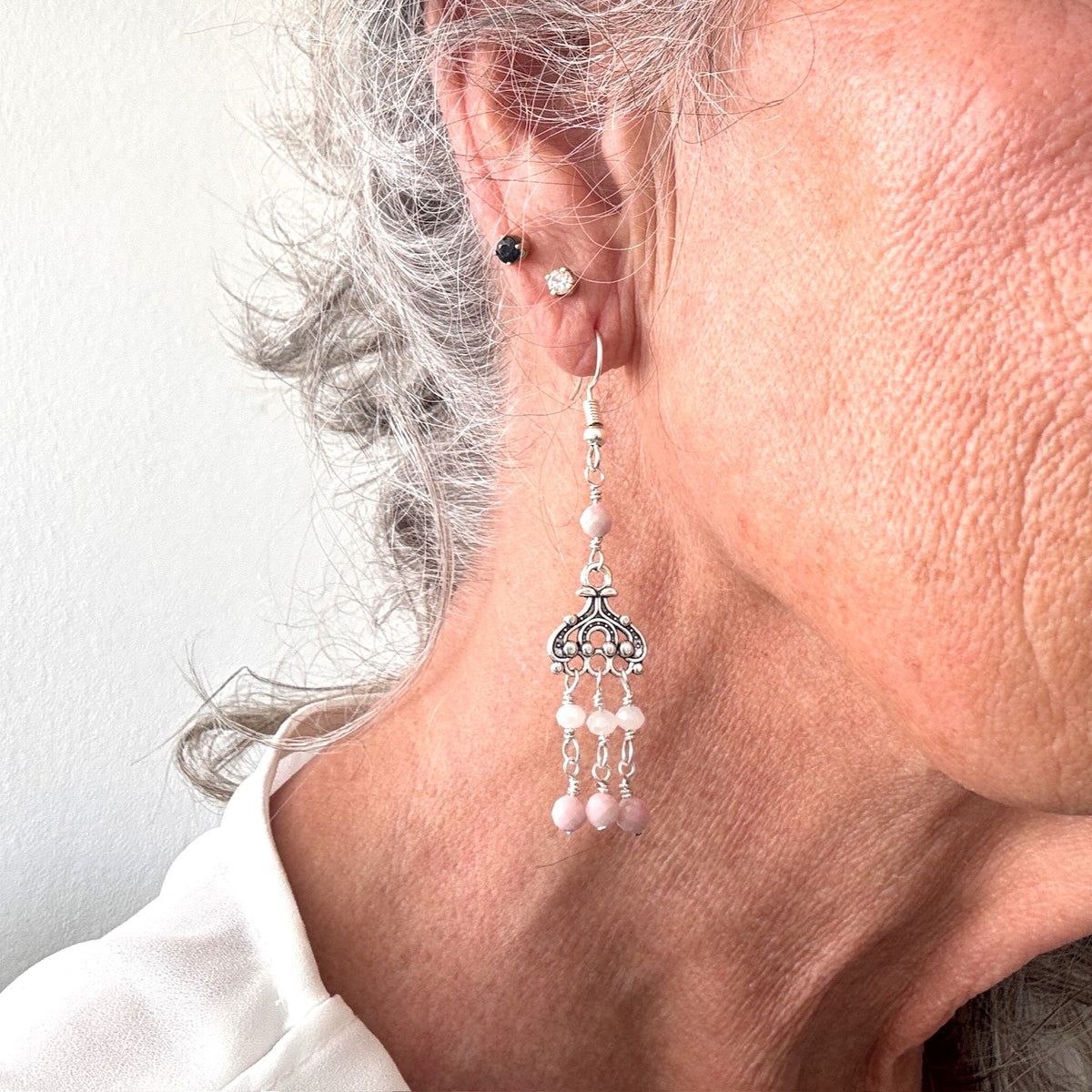 Pink opal chandelier earrings with ornate silver filigree and white crystal rondelle and pink opal bead dangles shown worn on model with silver hair wearing a white top
