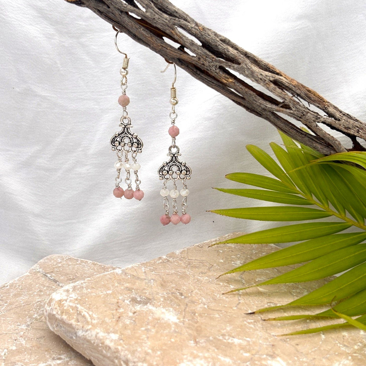 Pink opal earrings with ornate silver chandelier and white crystal rondelle and pink opal bead dangles hanging from driftwood against white linen background with palm frond and travertine
