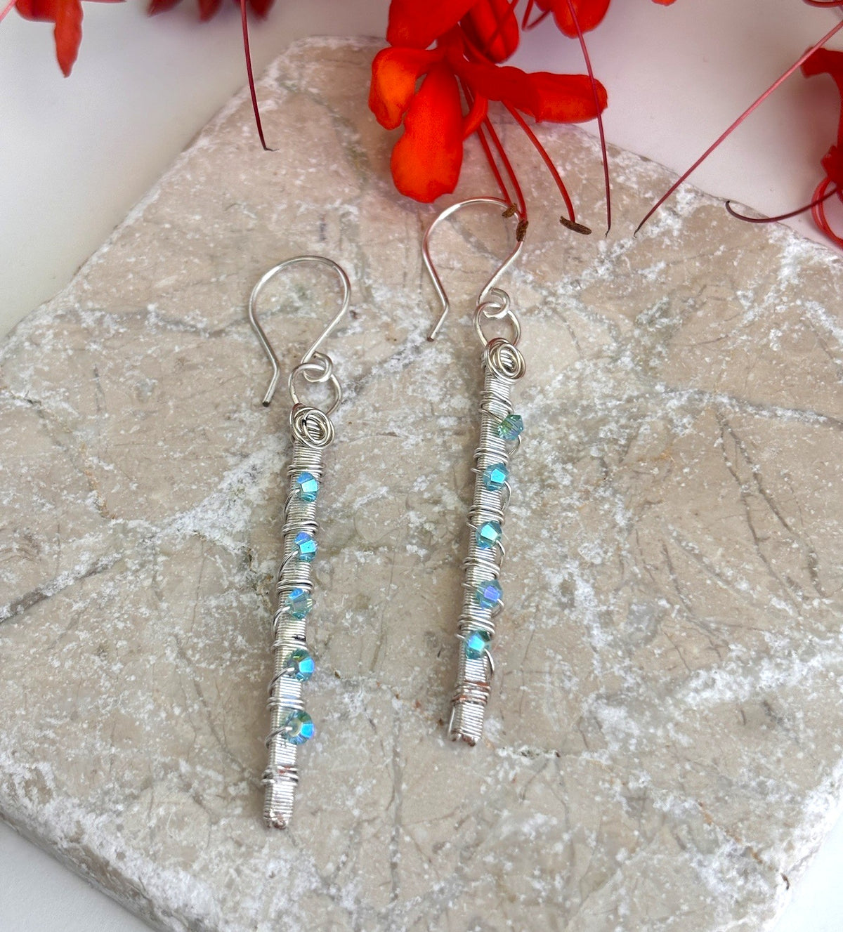 Handmade wire-wrapped aqua Swarovski crystal bar earrings with hand-coiled silver-plate ring displayed on granite surface with red tropical flowers