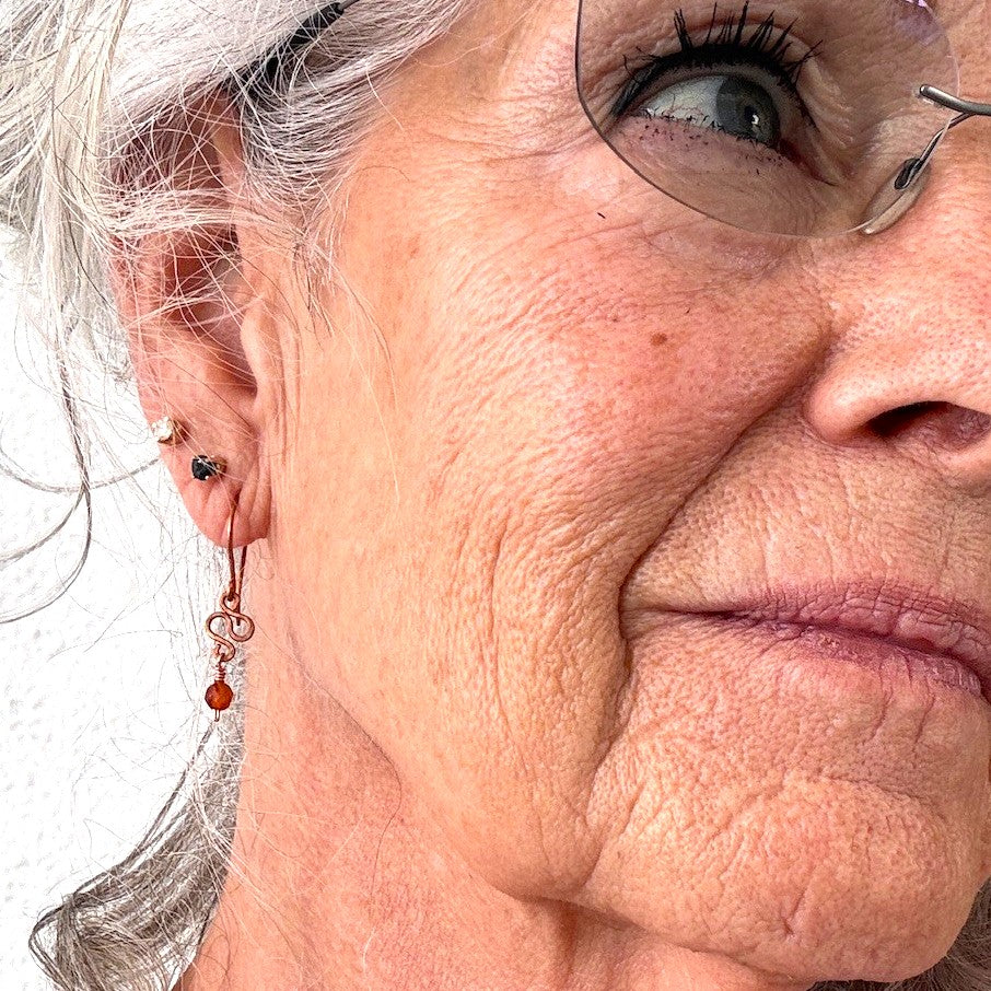 Carnelian earrings with hammered antique copper S-curve and faceted carnelian bead drop shown worn on model with silver hair and glasses