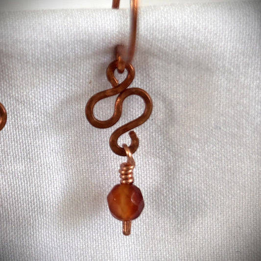 Close-up of handmade carnelian earring showing hand-formed hammered antique copper S-curve detail and wire-wrapped 4mm faceted carnelian bead drop on white linen