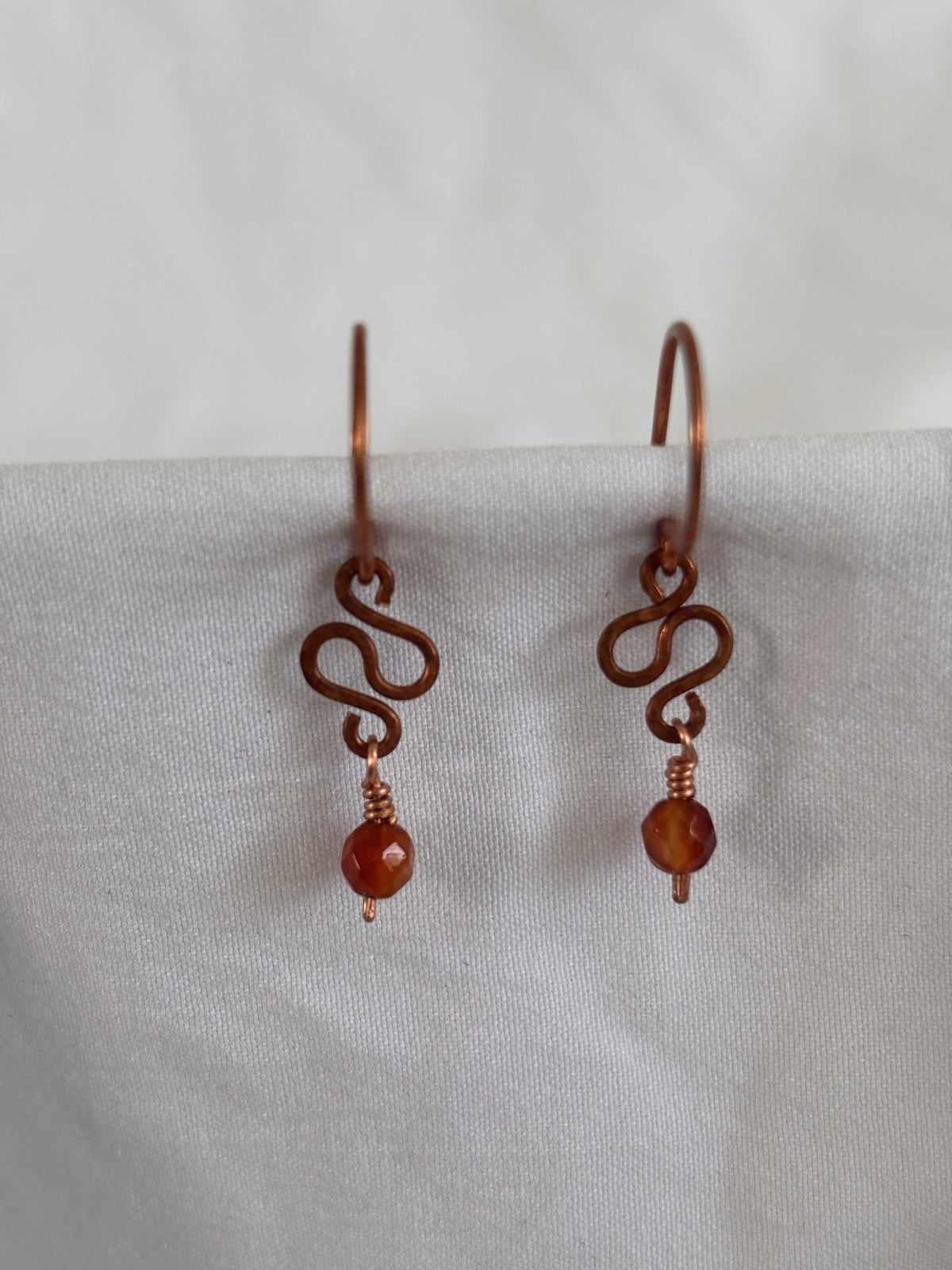 Handmade carnelian earrings with hammered antique copper ear wires, hand-formed S-curve, and wire-wrapped faceted carnelian bead drop, displayed against white linen background