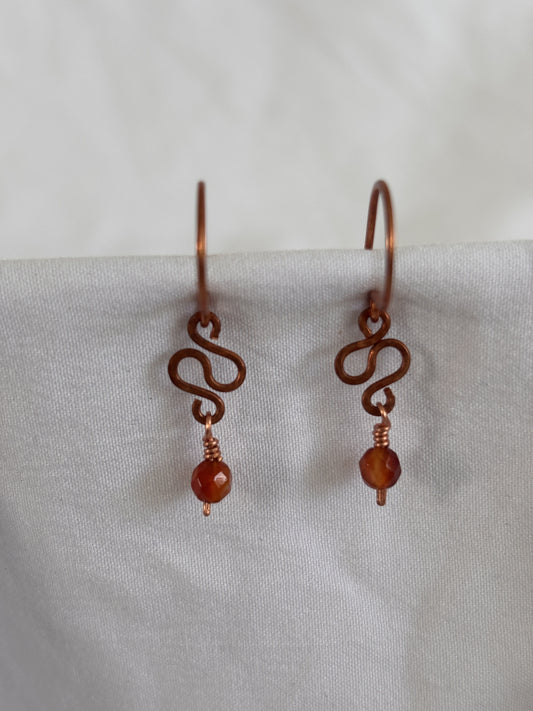 Handmade carnelian earrings with hammered antique copper ear wires, hand-formed S-curve, and wire-wrapped faceted carnelian bead drop, displayed against white linen background