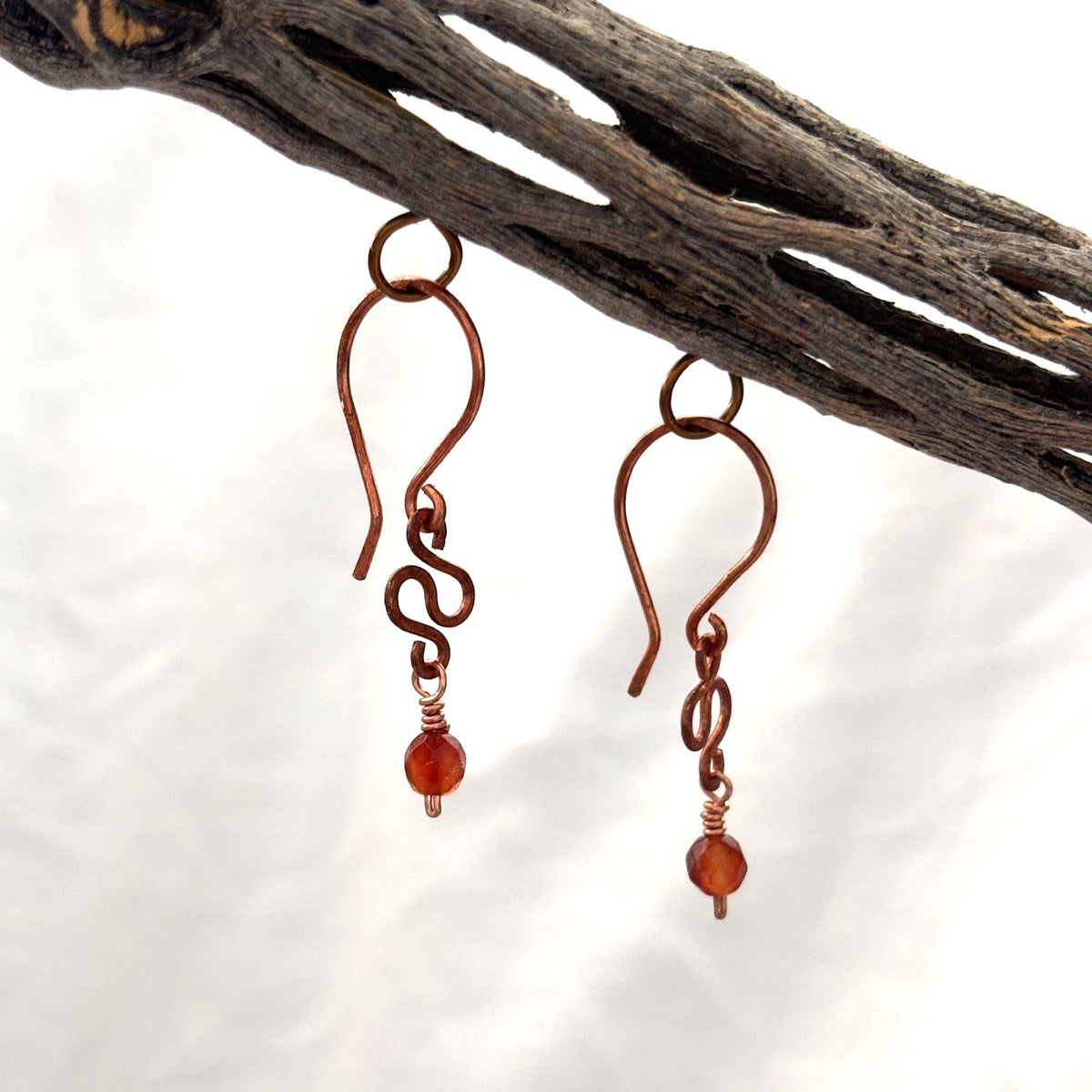 Handmade antique copper carnelian earrings with hammered S-curve and faceted carnelian bead drop shown in profile hanging from driftwood against white background