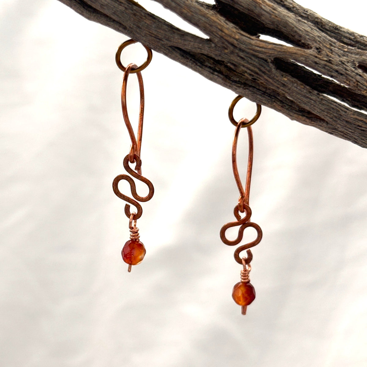 Carnelian earrings with hammered antique copper ear wire, hand-formed S-curve, and faceted carnelian bead drop hanging from driftwood against white background