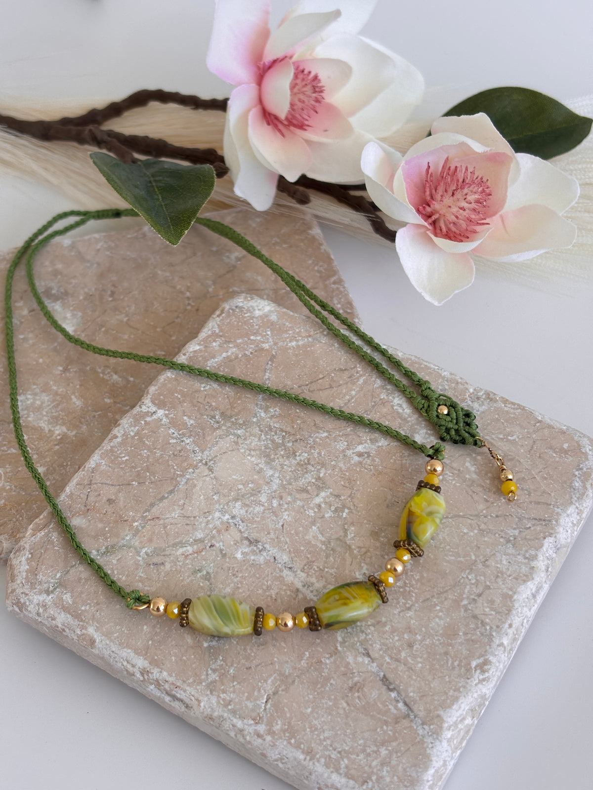 Handcrafted grass green macrame cord necklace with three twisted rectangle chartreuse lampwork glass beads, 4mm round gold-tone beads, 3mm opaque yellow crystal accents, and brass rondelle spacers, displayed on travertine tile with pink magnolia flowers and green leaf.
